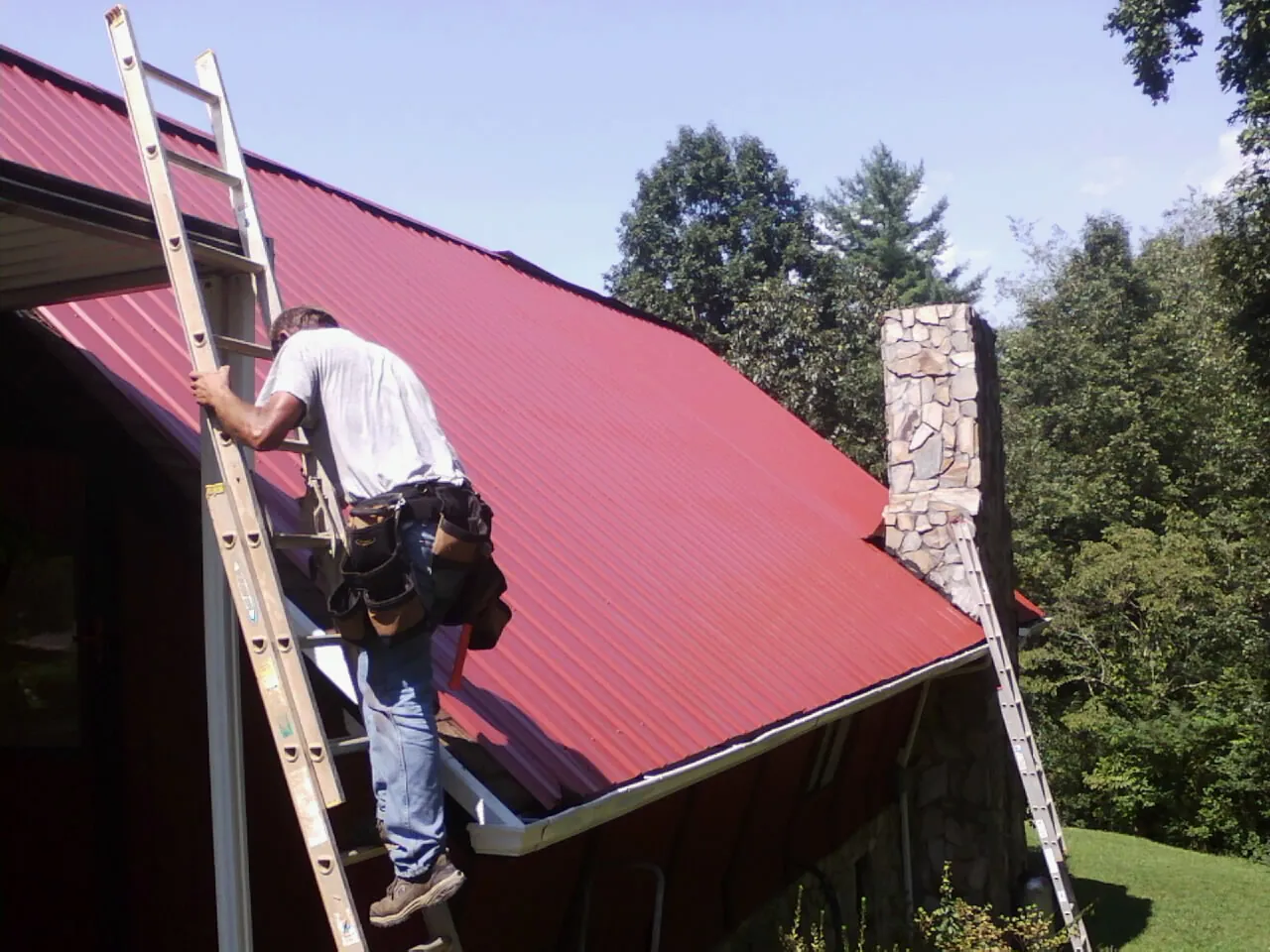 Roofer on a ladder beside a red metal roof and stone chimney — Soffit & Fascia Repair by Roofing Brothers in Kingsley